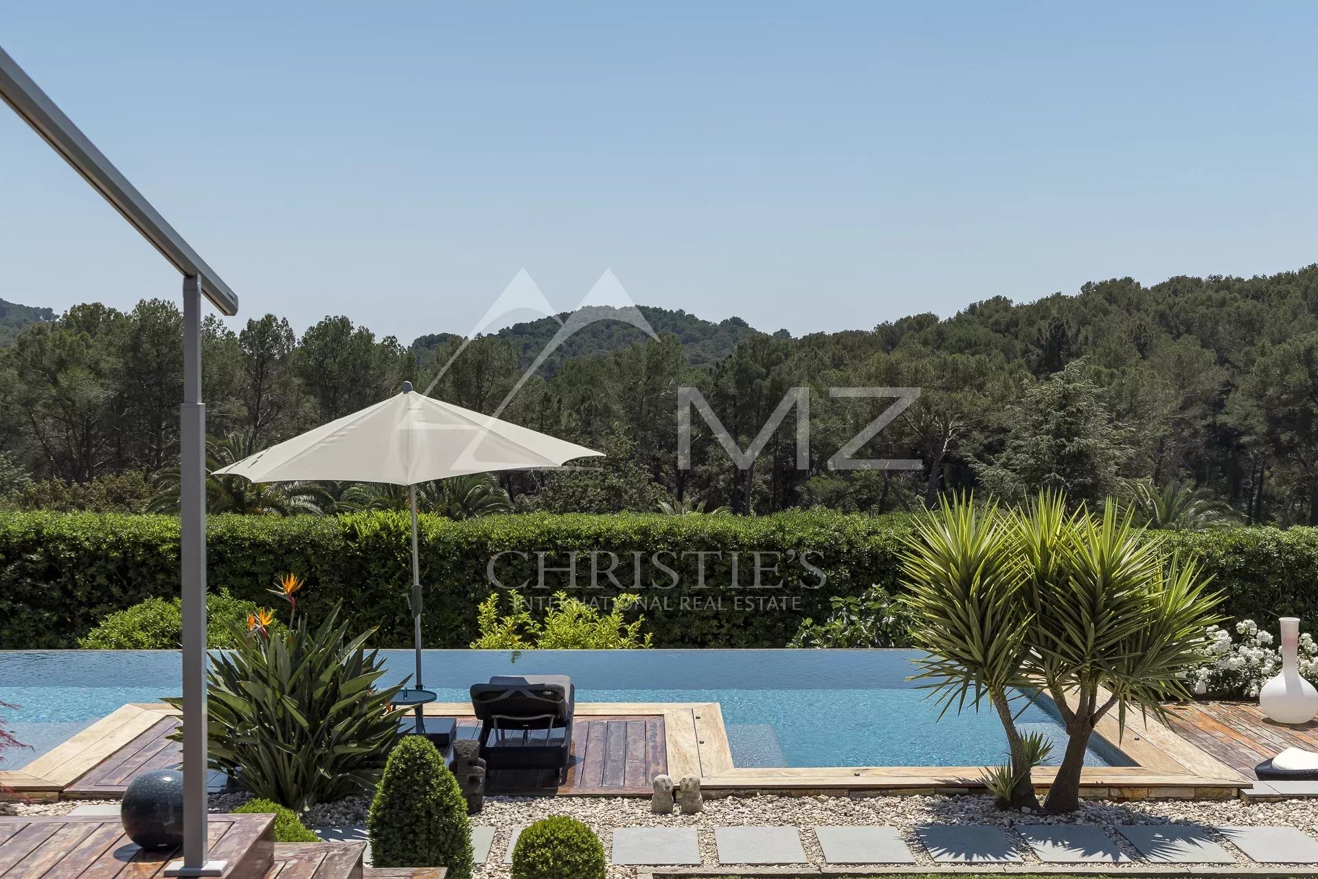 Infinity pool with a wooden deck, white umbrella and lounge chairs, surrounded by hedges and tropical plants with hills in the background.