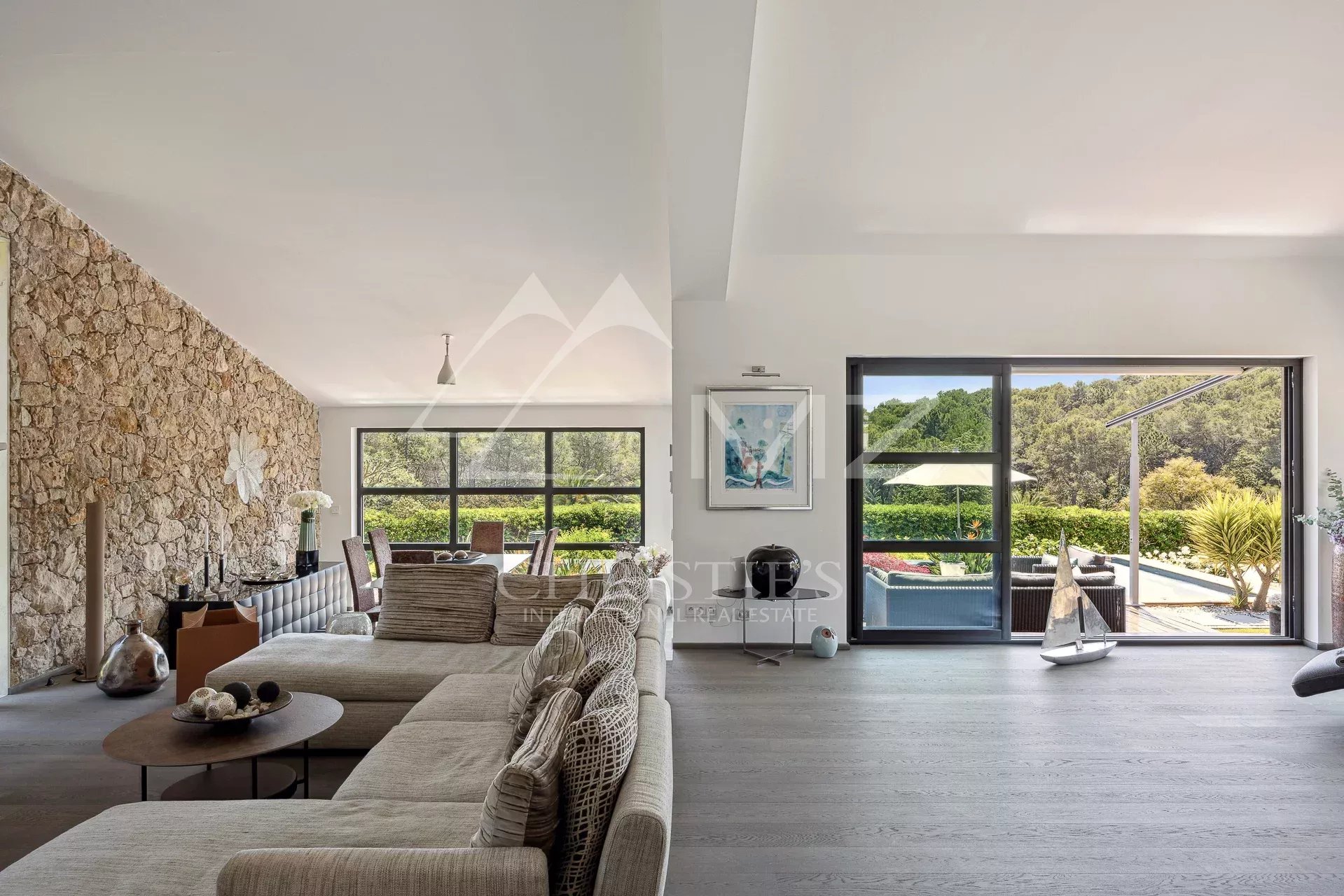 Bright open-plan living room with a stone feature wall, neutral sofa, and a views of the outdoor pool through large glass doors.