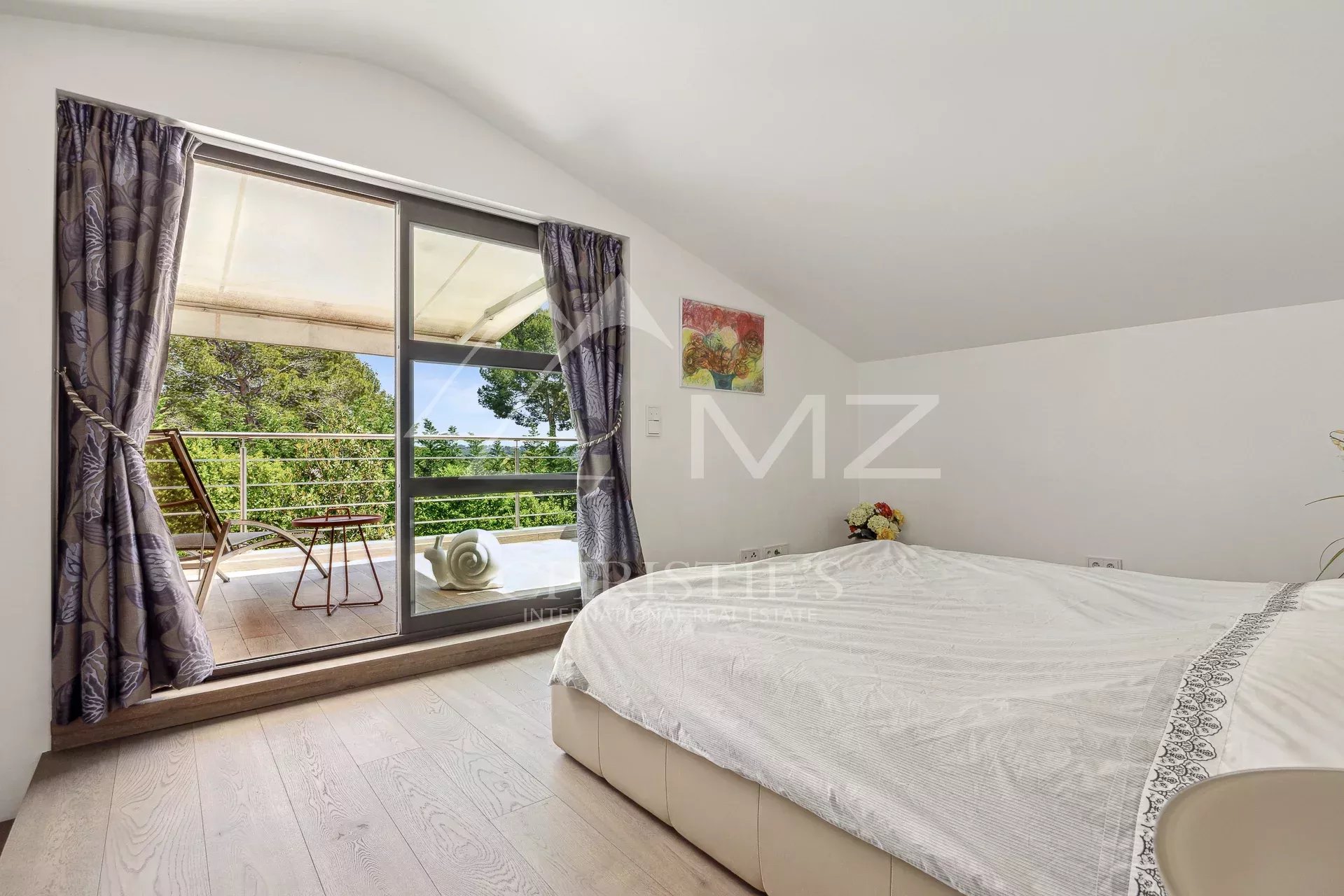 Bedroom with a large glass door to a balcony, purple patterned curtains, white bed, and wooden floor.