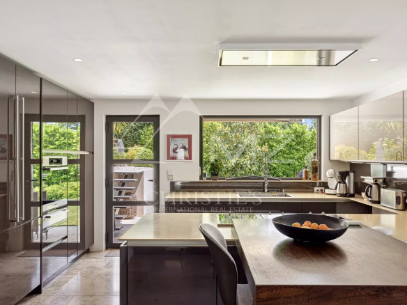 Modern kitchen with glossy gray cabinets, stainless appliances, and a wood island topped with a bowl of fruit, open to a sunny garden view.