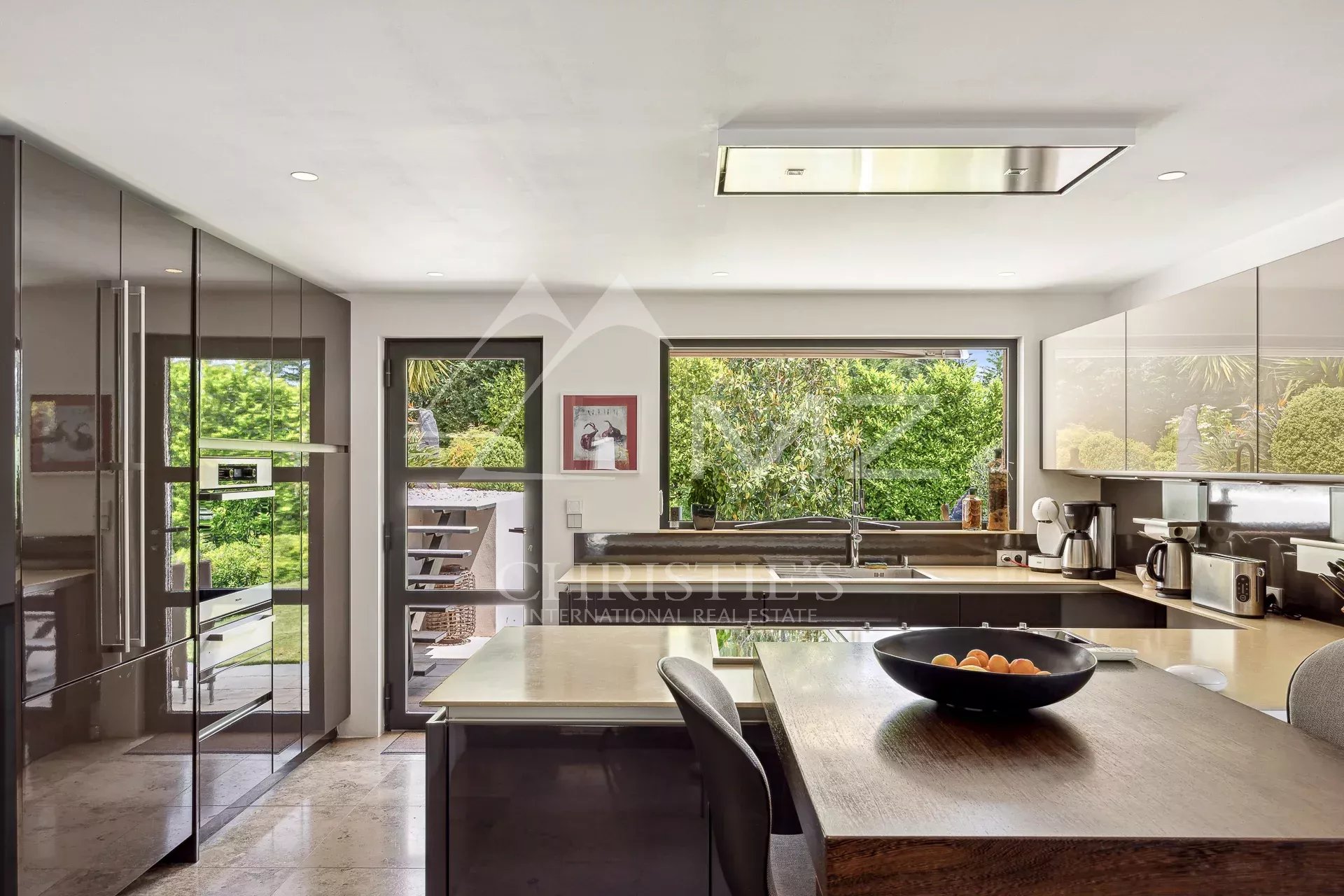 Modern kitchen with glossy gray cabinets, stainless appliances, and a wood island topped with a bowl of fruit, open to a sunny garden view.