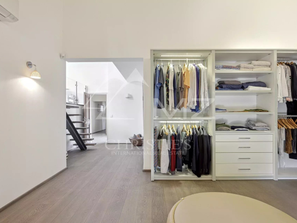 Open walk-in closet with white shelving, hanging clothes, folded sweaters, and drawers; adjacent hallway and stairs visible in a bright, modern space.