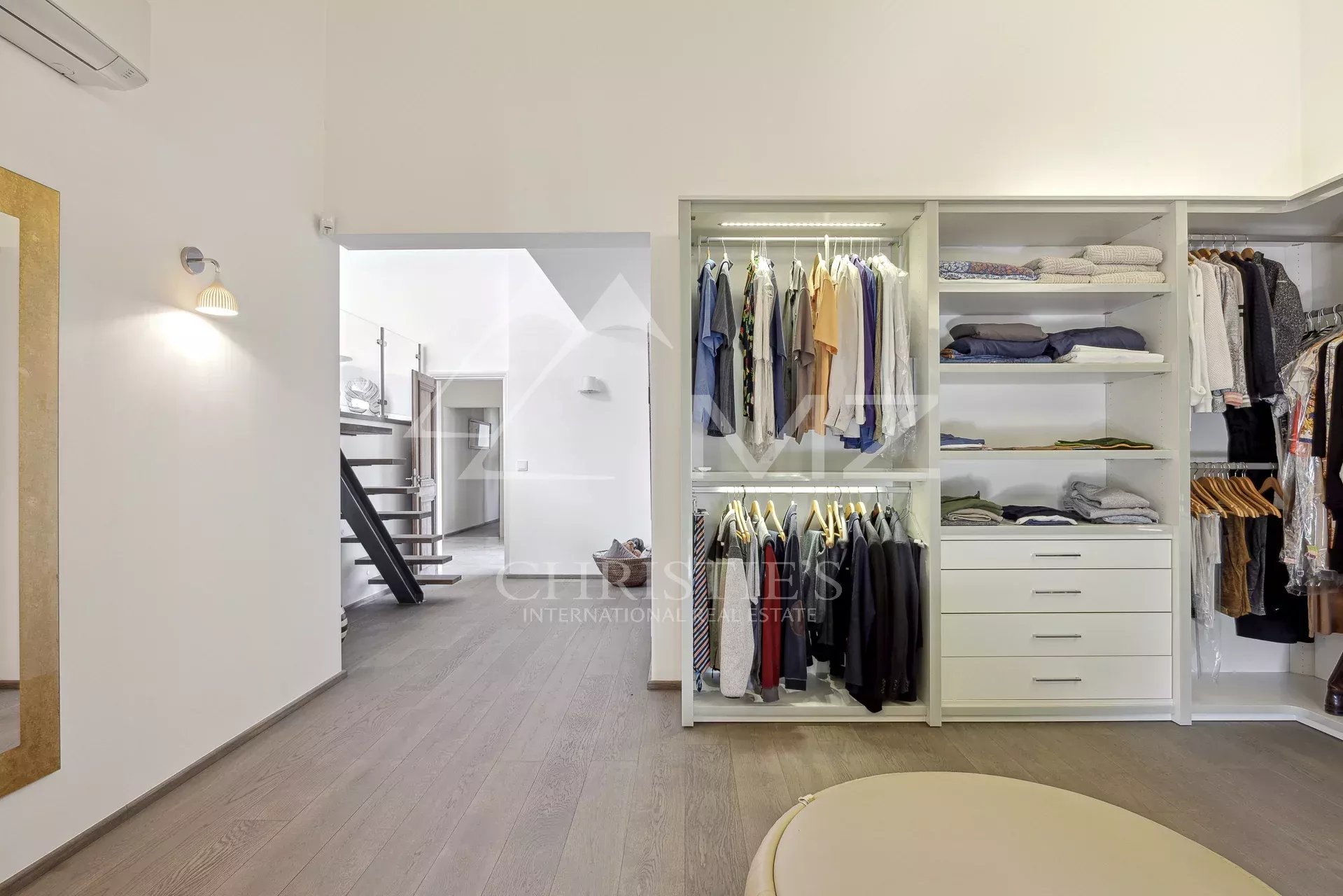 Open walk-in closet with white shelving, hanging clothes, folded sweaters, and drawers; adjacent hallway and stairs visible in a bright, modern space.