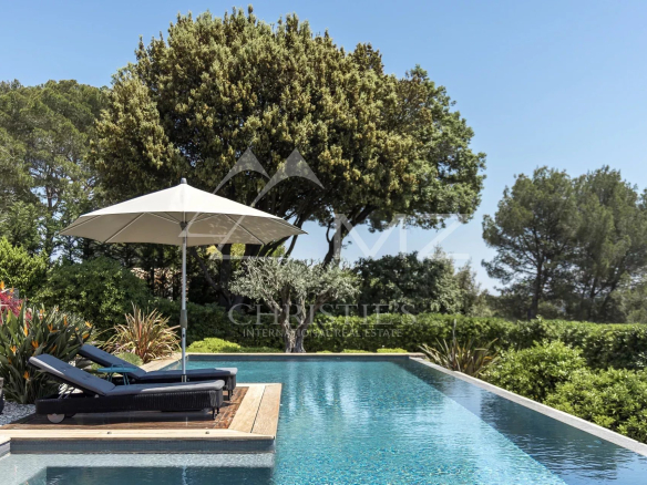 Pool deck with two black lounge chairs under a large white parasol, surrounded by trees on a sunny day.