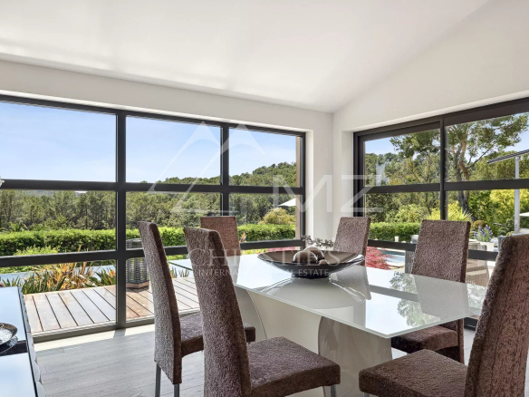 Bright dining room with a glass table and six brown upholstered chairs, large windows showing a green landscape outside.