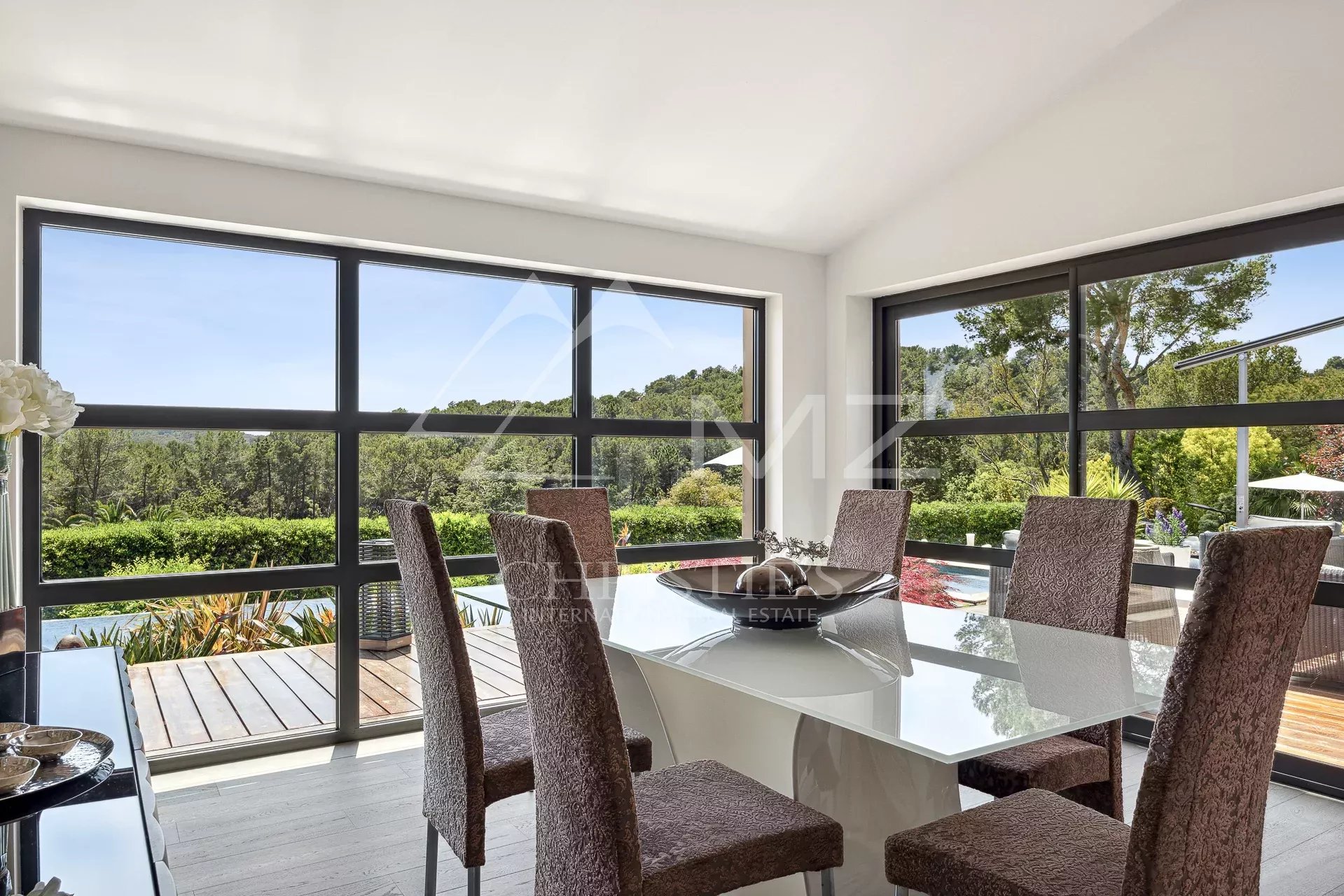 Bright dining room with a glass table and six brown upholstered chairs, large windows showing a green landscape outside.