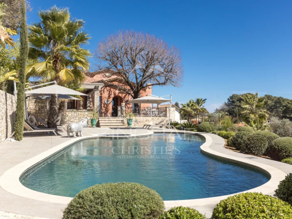 Curved swimming pool on a sunny terrace with stone walls, palm trees, and a peach-colored villa in the background.