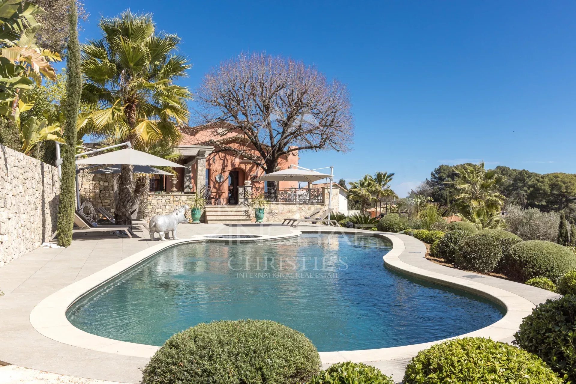 Curved swimming pool on a sunny terrace with stone walls, palm trees, and a peach-colored villa in the background.