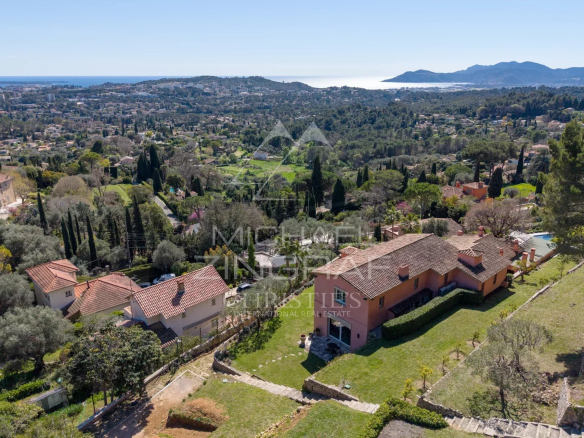 Aerial view of a hillside neighborhood with terracotta-roofed houses, green gardens, and distant coastline.