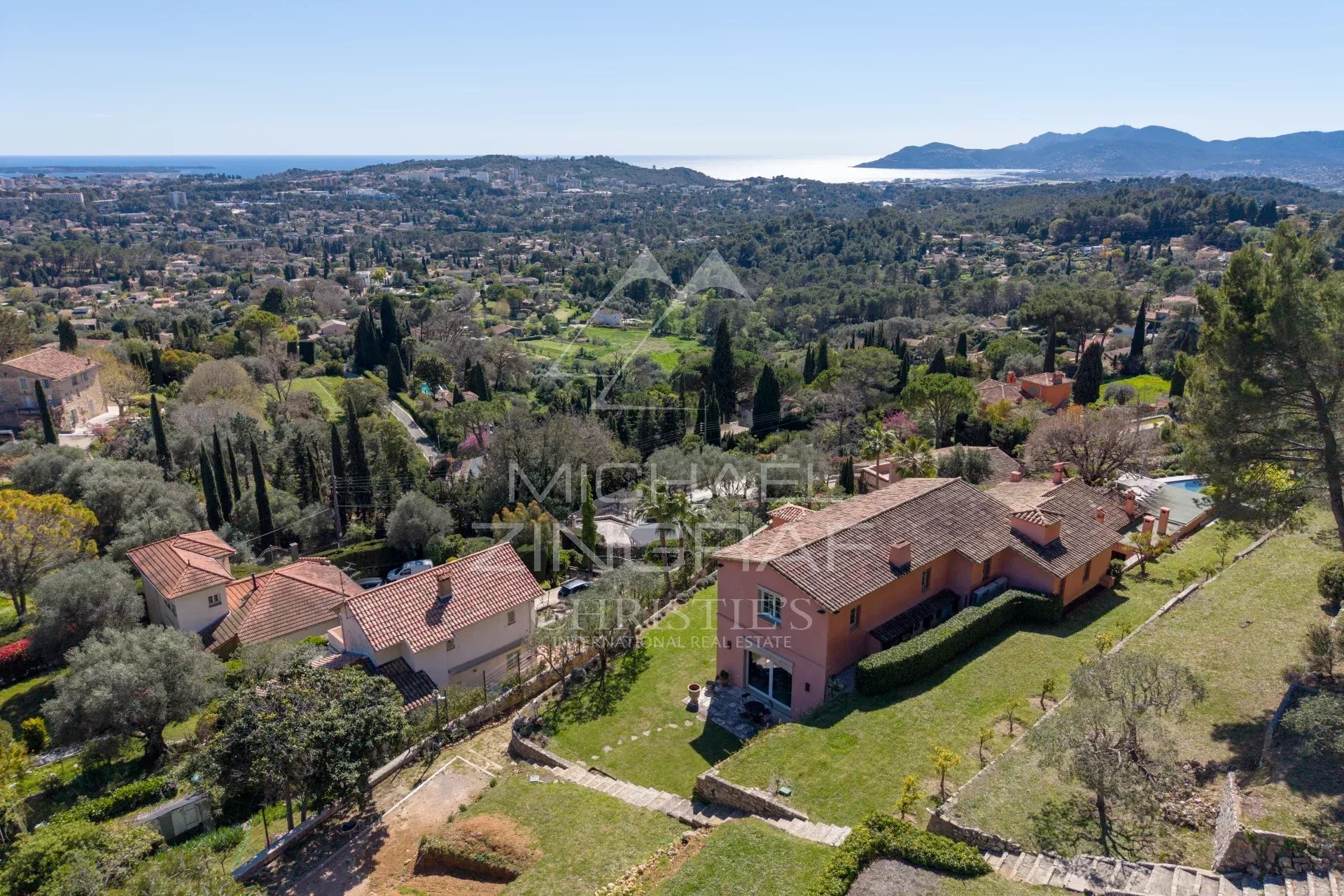 Aerial view of a hillside neighborhood with terracotta-roofed houses, green gardens, and distant coastline.