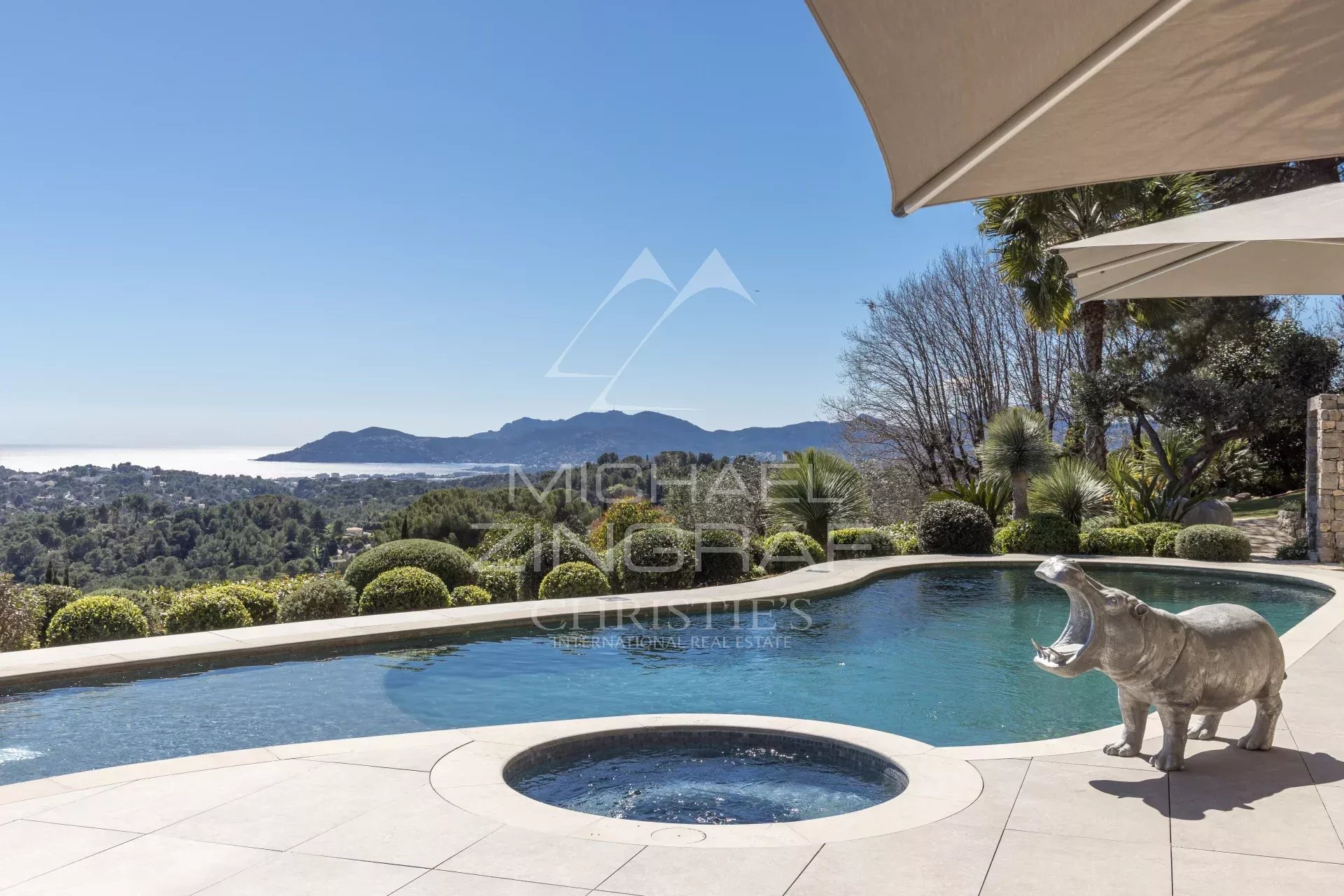 Luxurious pool terrace with a circular hot tub, beige tiles, and a coastal valley view; a hippo statue by the pool.