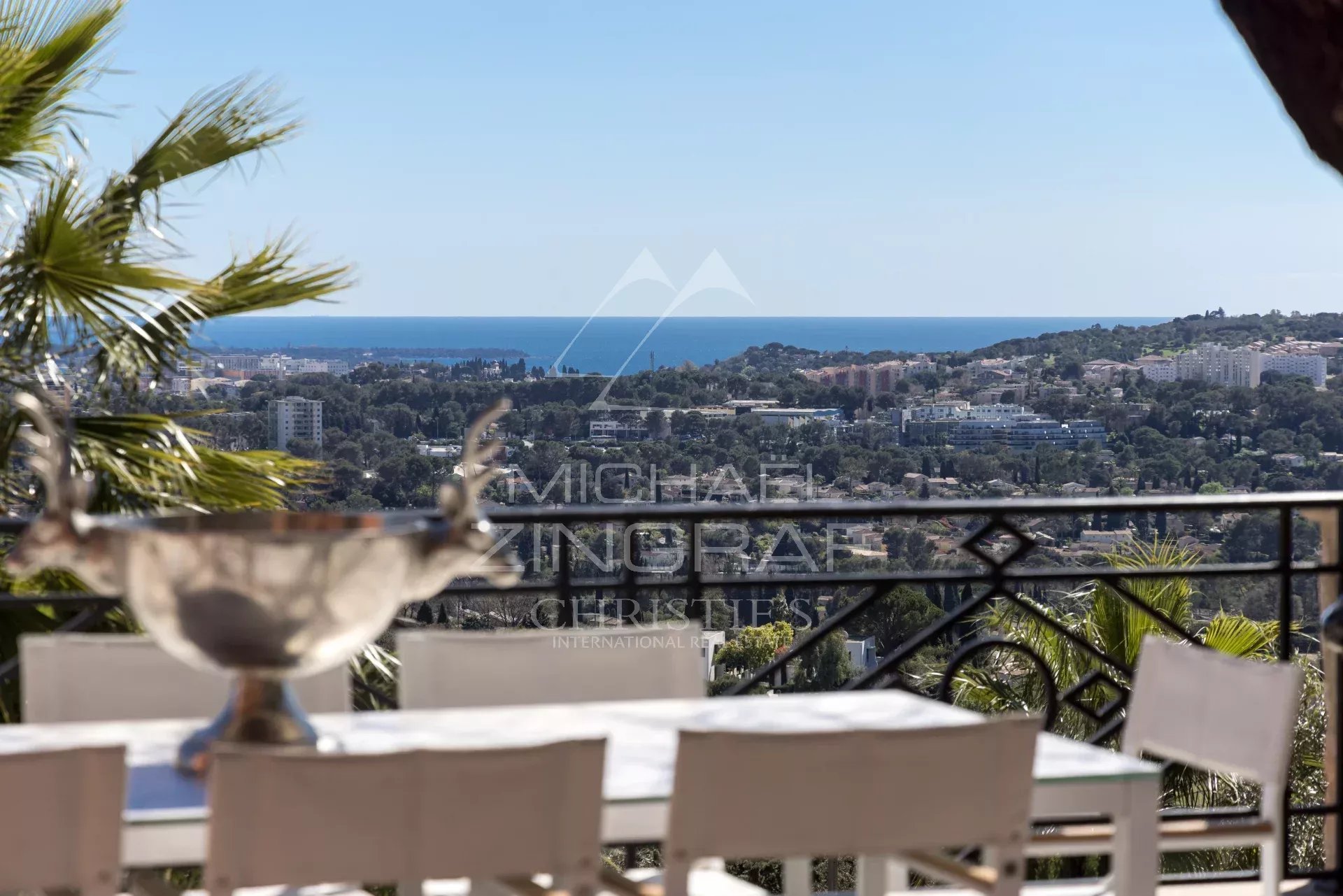 Terrace with a white dining table and chairs, palm fronds on the left, railing, and a panoramic view of the sea and coastline beyond.