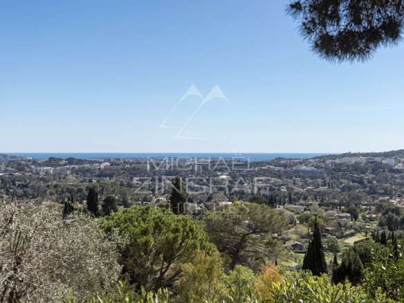 Panoramic hillside view over a coastal town with blue sea on the horizon and green trees in the foreground under a clear sky