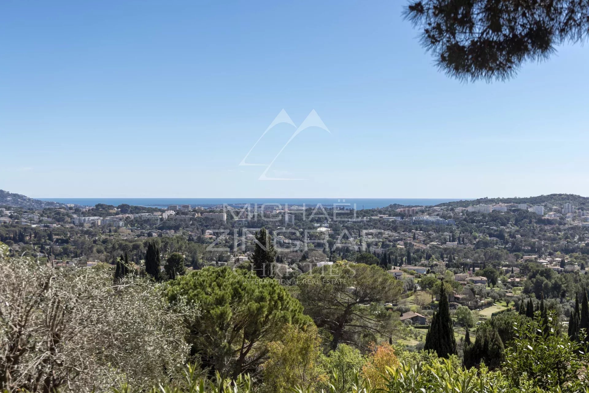Panoramic hillside view over a coastal town with blue sea on the horizon and green trees in the foreground under a clear sky