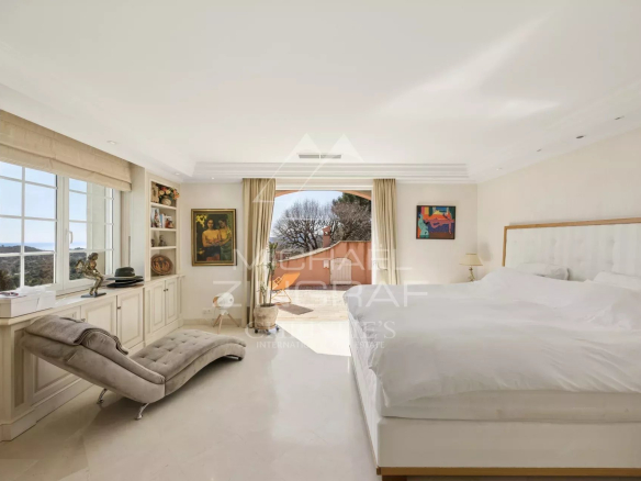 Bright luxury bedroom with a king bed, white headboard, a beige chaise lounge, built-in shelves, and a balcony overlooking trees.