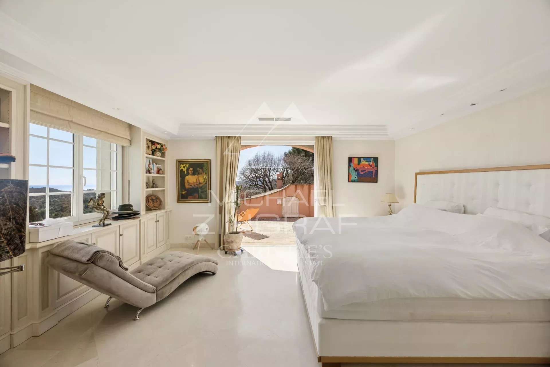 Bright luxury bedroom with a king bed, white headboard, a beige chaise lounge, built-in shelves, and a balcony overlooking trees.