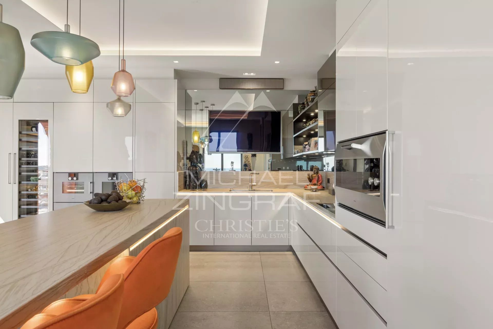 Modern white kitchen with a marble island, orange bar stools, and pendant lights hanging above the island.