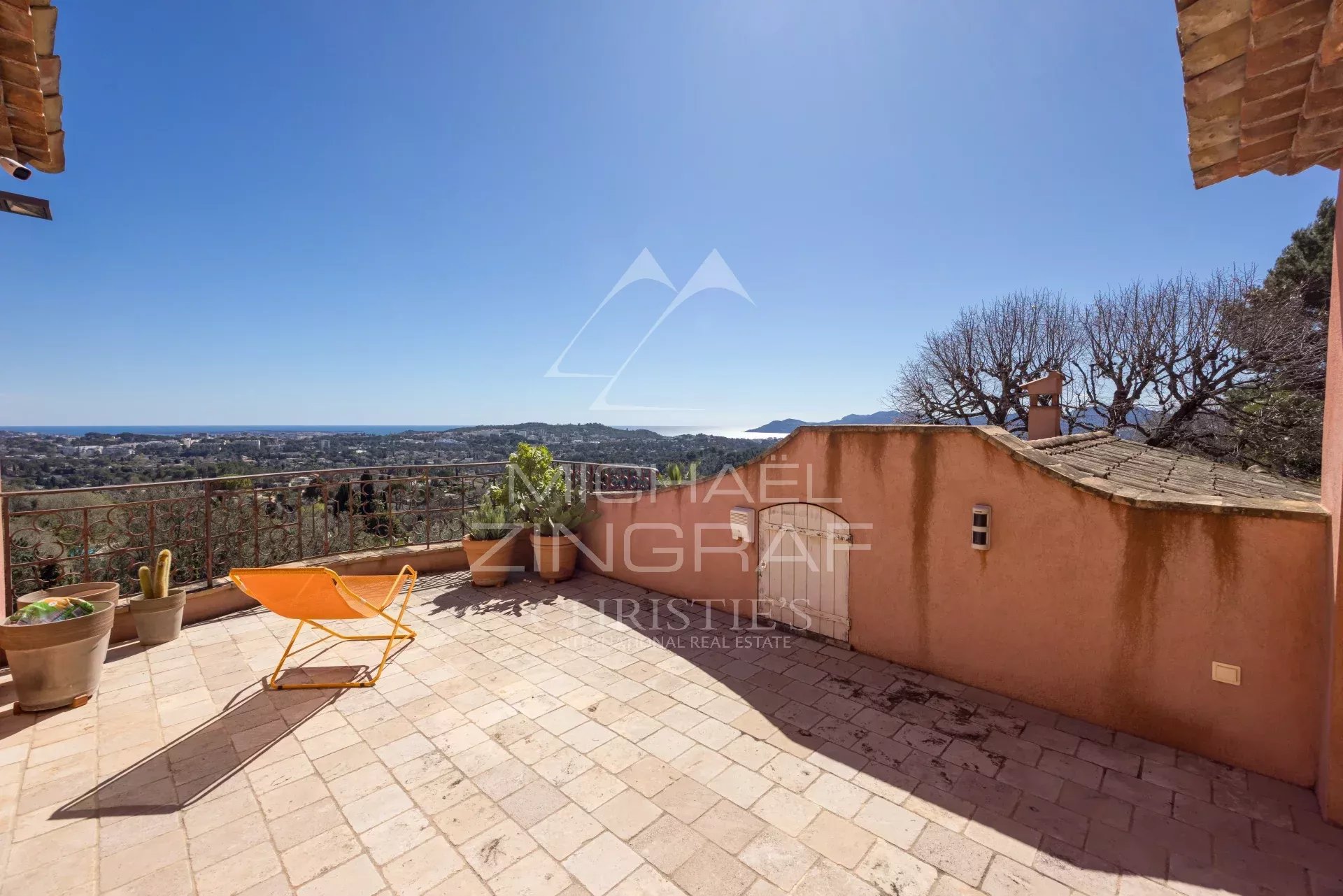 Terrace with a tiled floor, orange lounge chair, and potted plants, overlooking a coastal town and the sea under a clear blue sky.