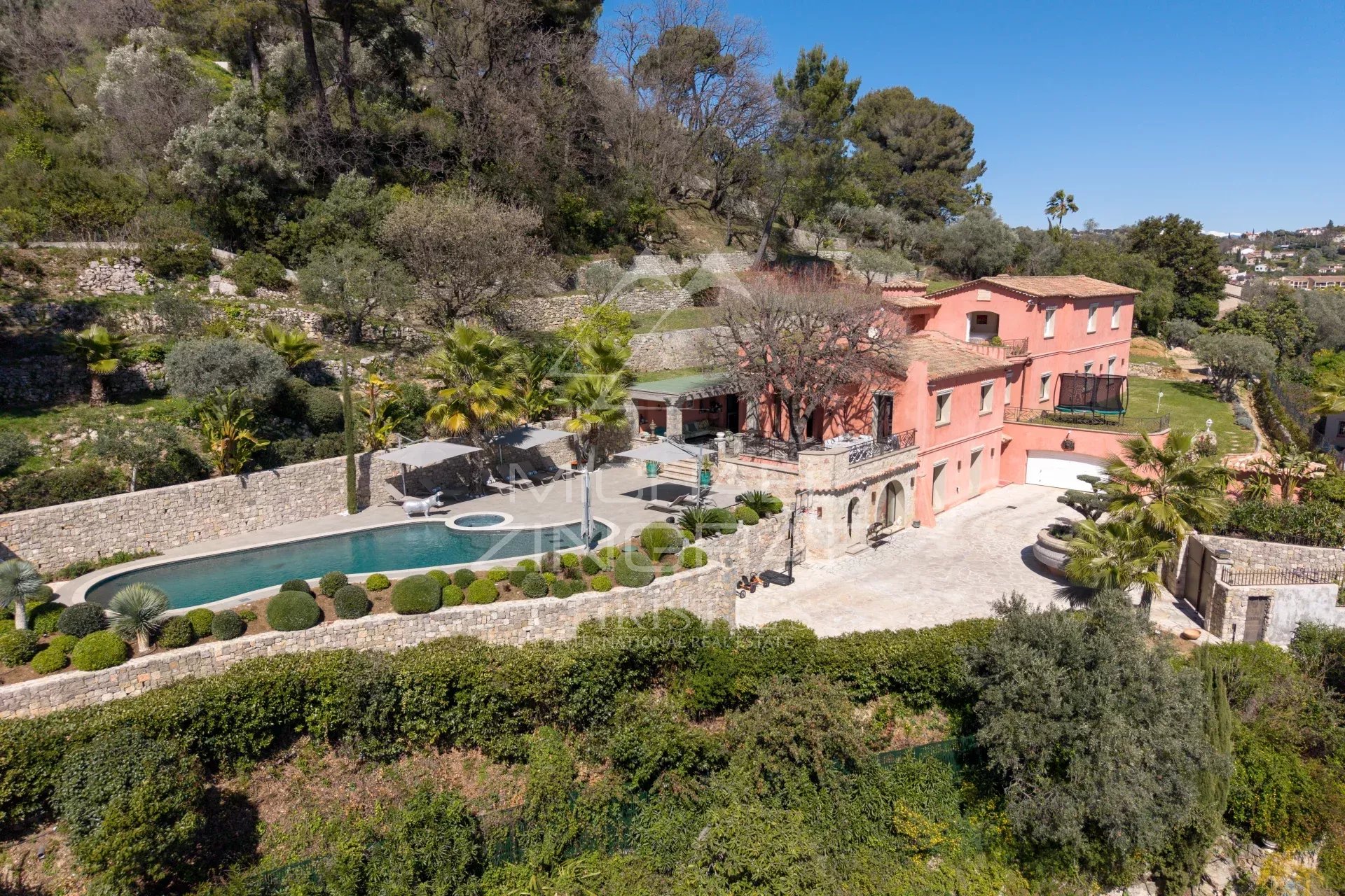A pink hillside villa with a curving pool, stone patio, and manicured shrubs overlooking terraced gardens.