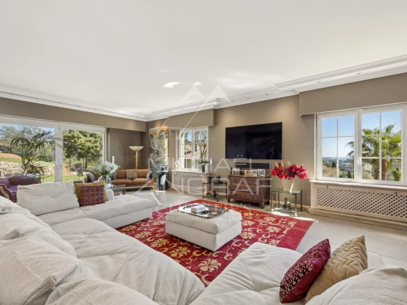 Spacious living room with a white sectional, red patterned rug, and a wall-mounted TV. Outdoors visible through large windows.