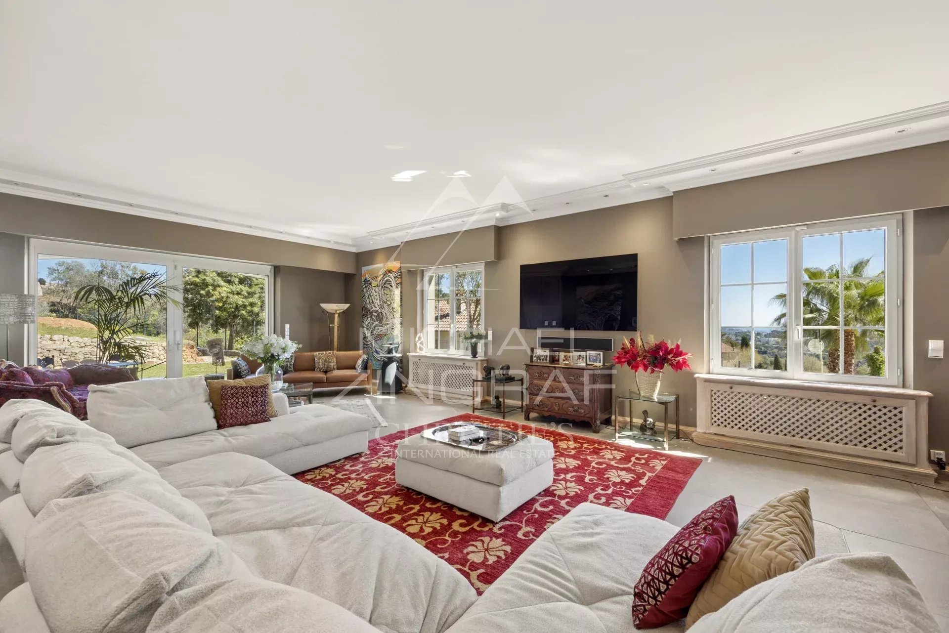 Spacious living room with a white sectional, red patterned rug, and a wall-mounted TV. Outdoors visible through large windows.