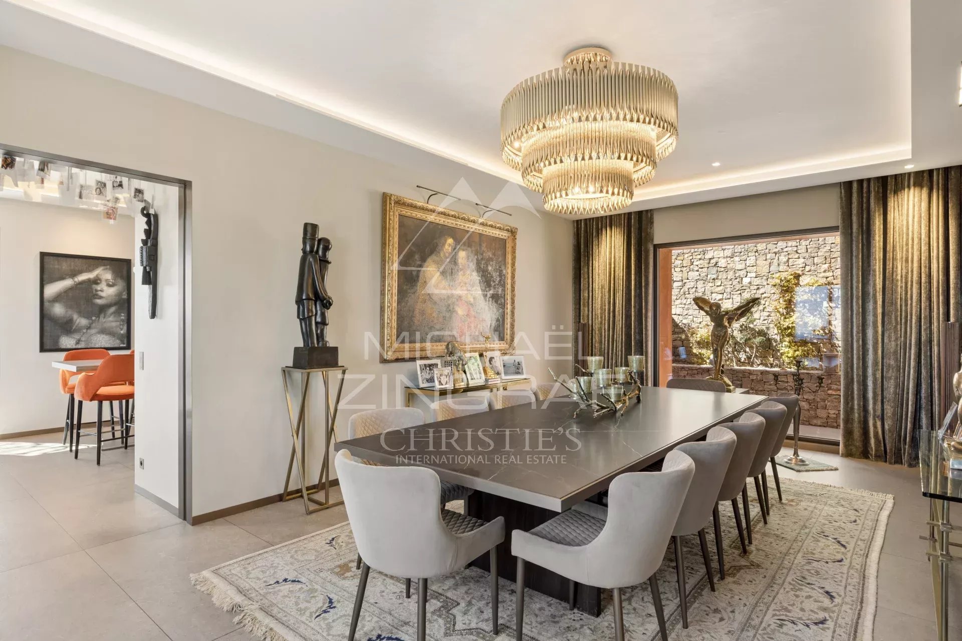 Luxurious dining room with a long glass-top table, upholstered chairs, and a tiered chandelier centerpiece.