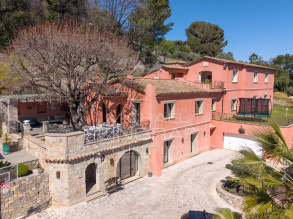 Pink Mediterranean villa with terracotta roof, stone terrace, and leafless tree in a sunny courtyard garden area, with outdoor seating and a trampoline on the lawn nearby.