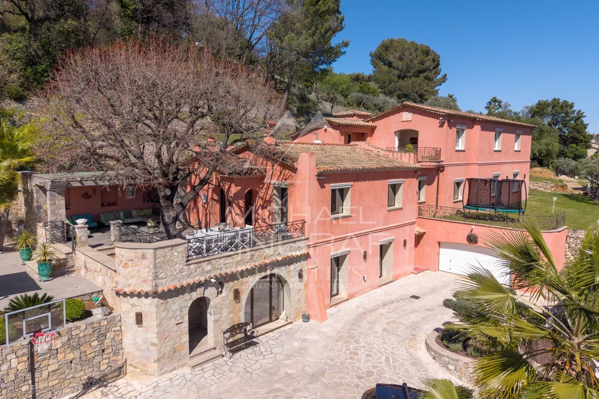 Pink Mediterranean villa with terracotta roof, stone terrace, and leafless tree in a sunny courtyard garden area, with outdoor seating and a trampoline on the lawn nearby.
