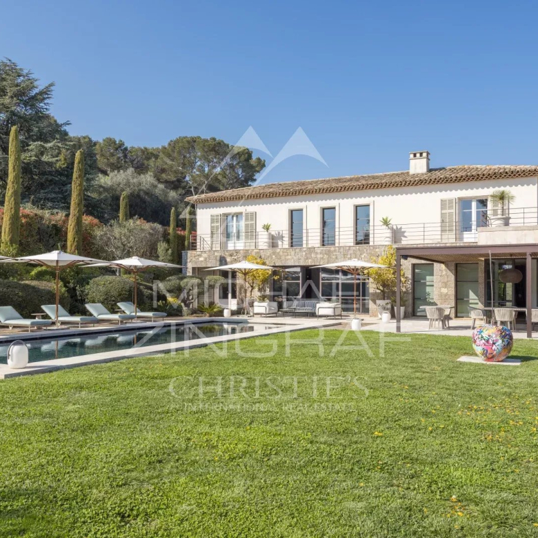 Villa with a rectangular pool, lounge chairs under umbrellas, and a spacious lawn in a sunny backyard. A two-story house with a terrace and outdoor dining area sits beyond the pool.