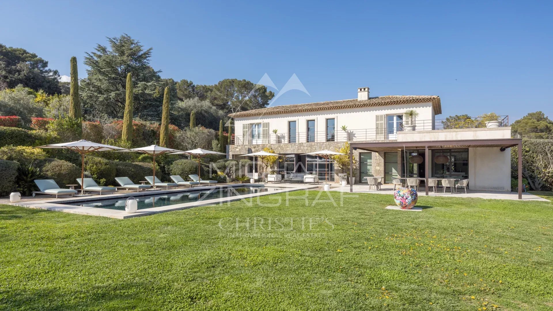 Villa with a rectangular pool, lounge chairs under umbrellas, and a spacious lawn in a sunny backyard. A two-story house with a terrace and outdoor dining area sits beyond the pool.