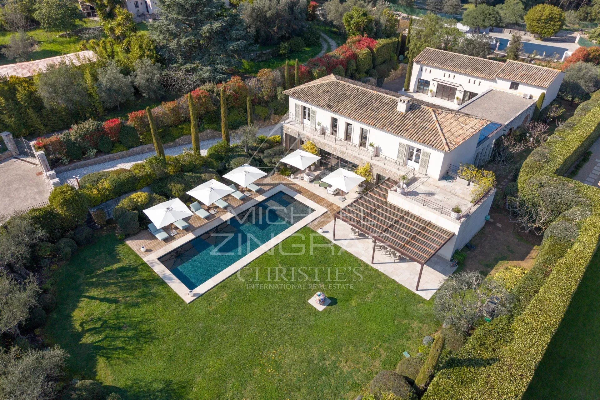 Aerial view of a white villa with a rectangular pool, poolside loungers and umbrellas, and a manicured garden yard.