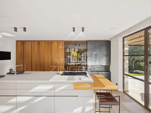 Modern kitchen with a white island, warm wood cabinets, and large glass doors opening to a garden.