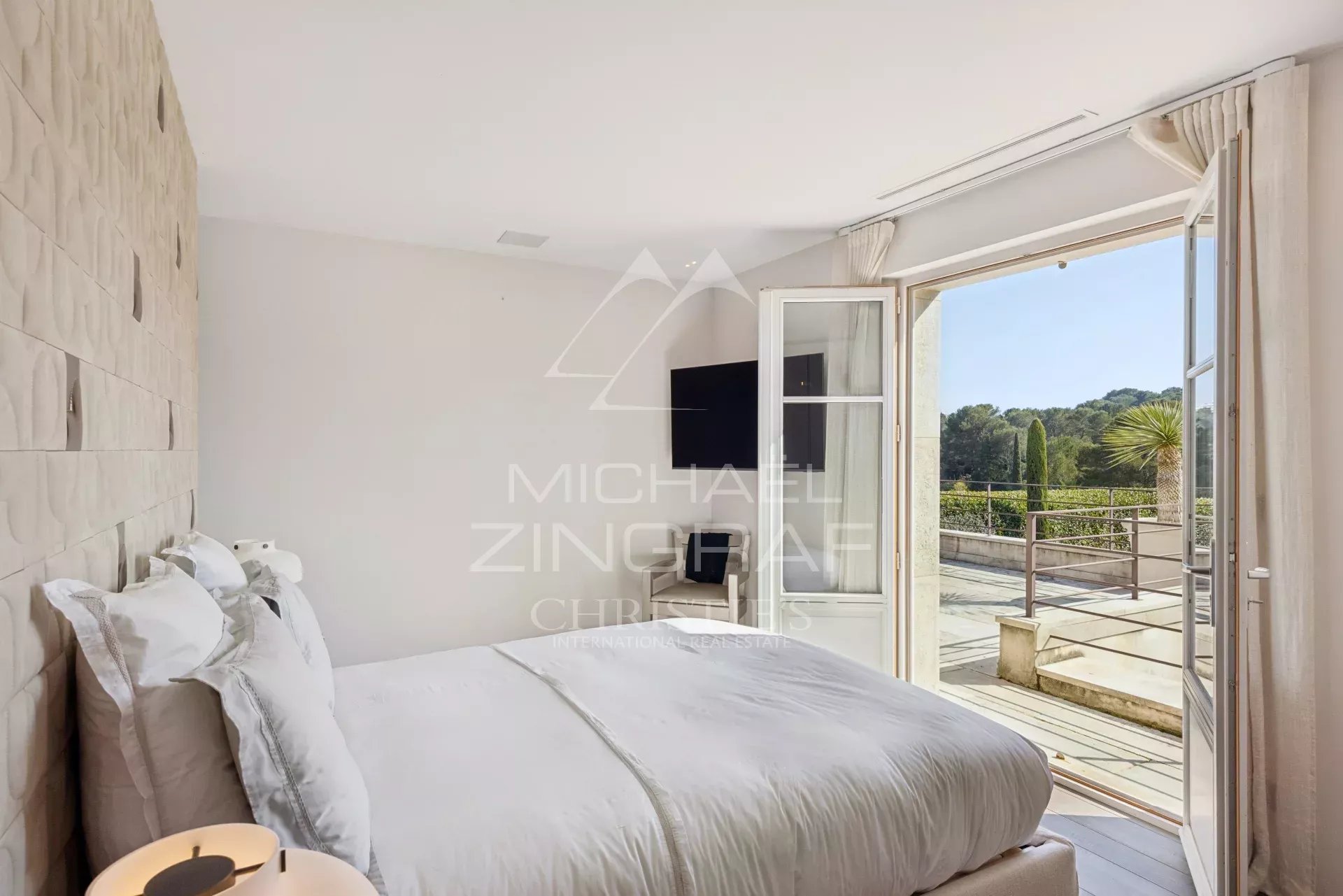 Bright white bedroom with a padded textured headboard, white bedding, and a wall-mounted TV near the windowed doors to a balcony with a garden view