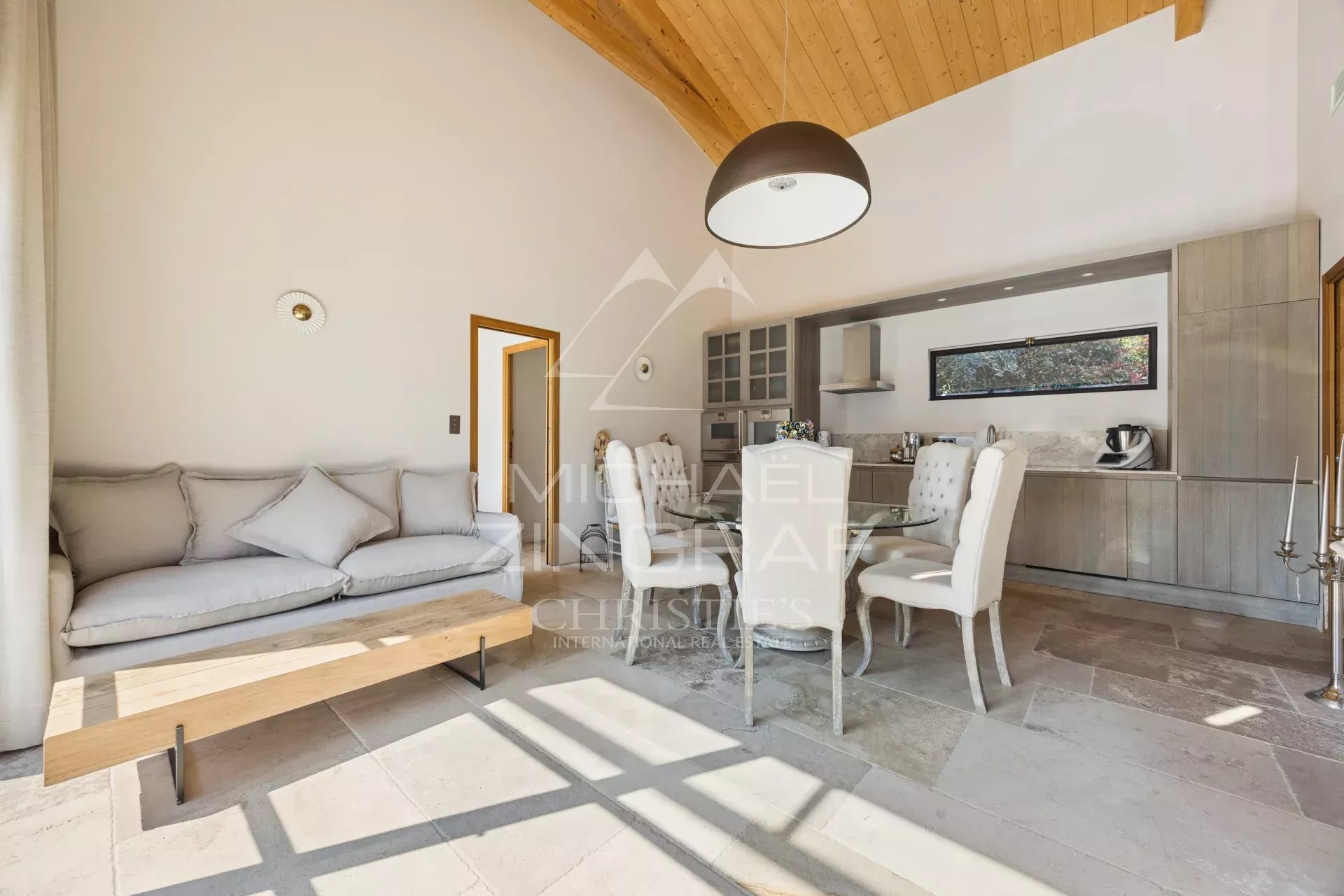 Open-plan living and dining area with light gray sofa, wooden coffee table, and white upholstered dining chairs facing a modern kitchen.