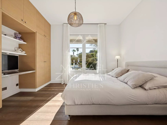 Bright bedroom with built-in wooden wardrobe, low upholstered bed, and a window with white curtains letting in sunlight.