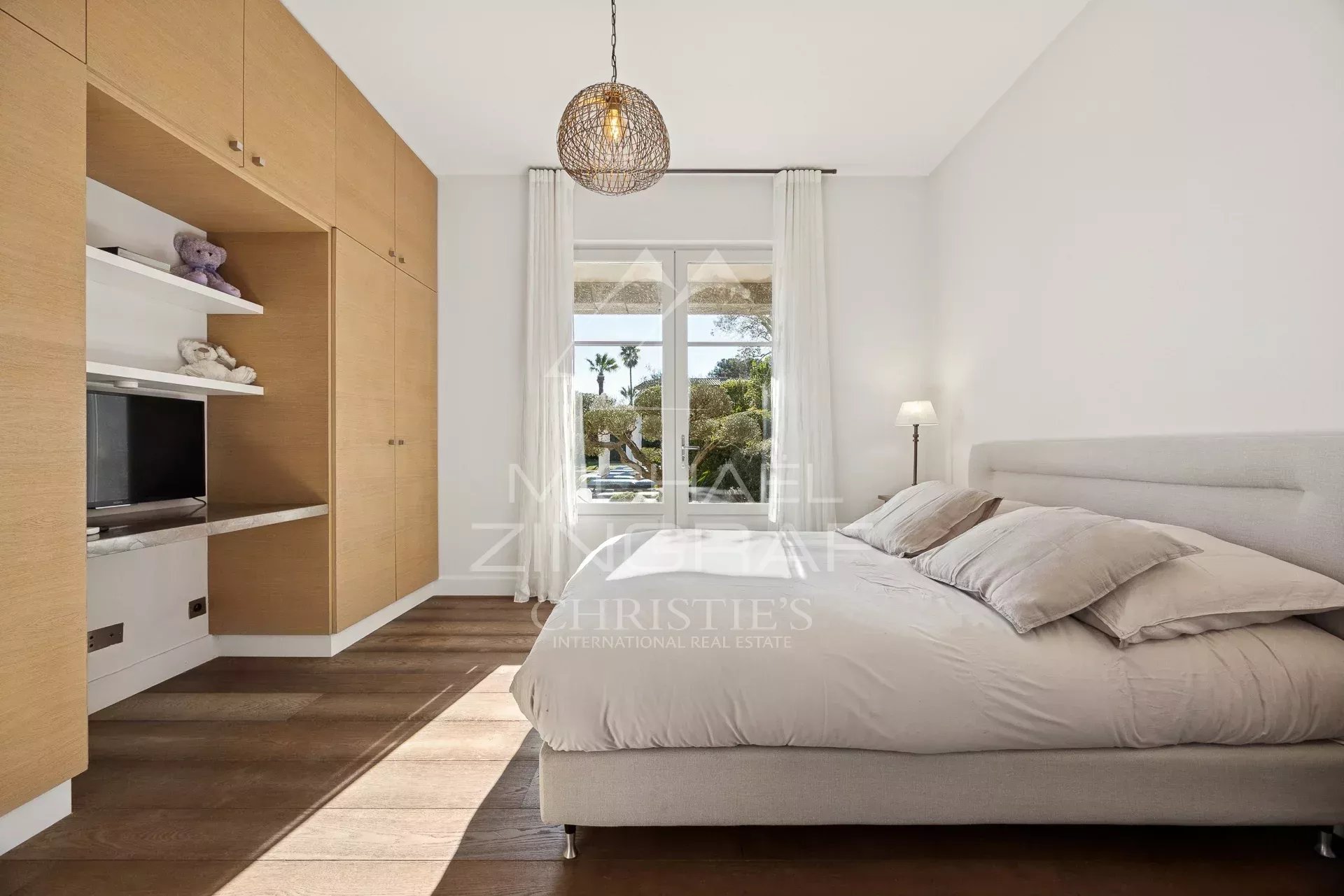 Bright bedroom with built-in wooden wardrobe, low upholstered bed, and a window with white curtains letting in sunlight.