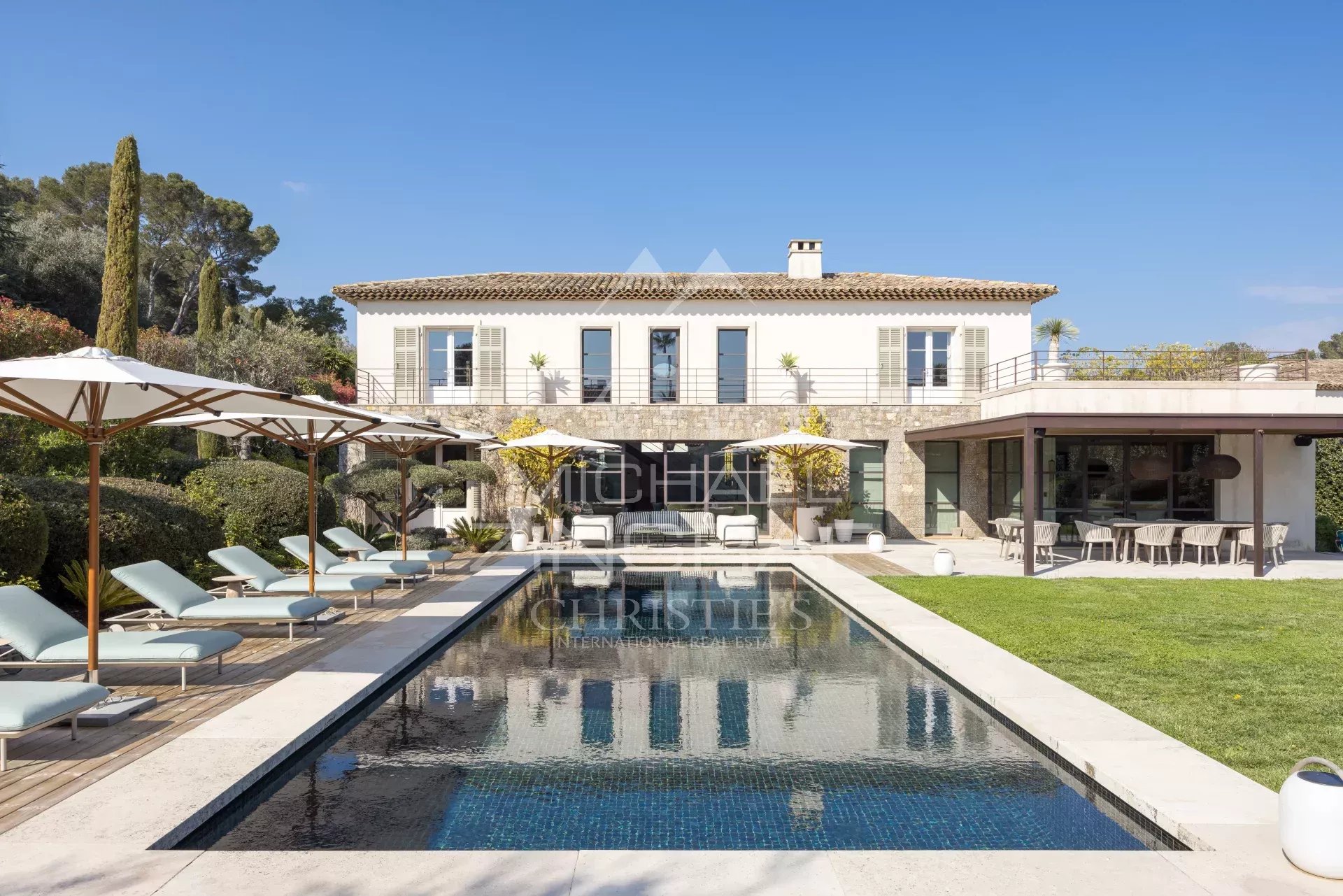 Luxurious two-story villa with a stone terrace, surrounding pool, lounge chairs, and white umbrellas on a sunny day.