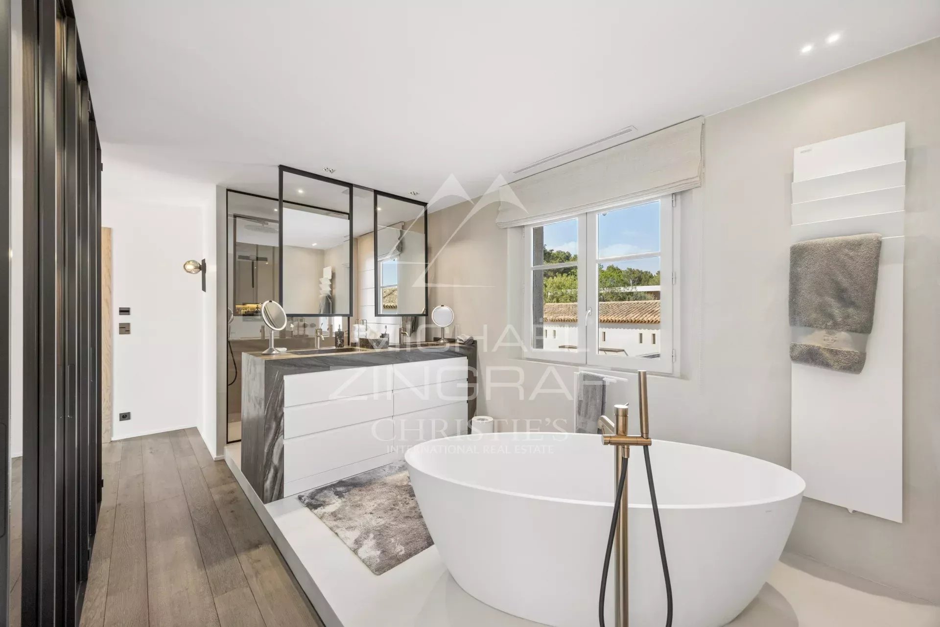 Contemporary bathroom with a freestanding white tub, marble-topped vanity, and a glass-framed partition by a large window.