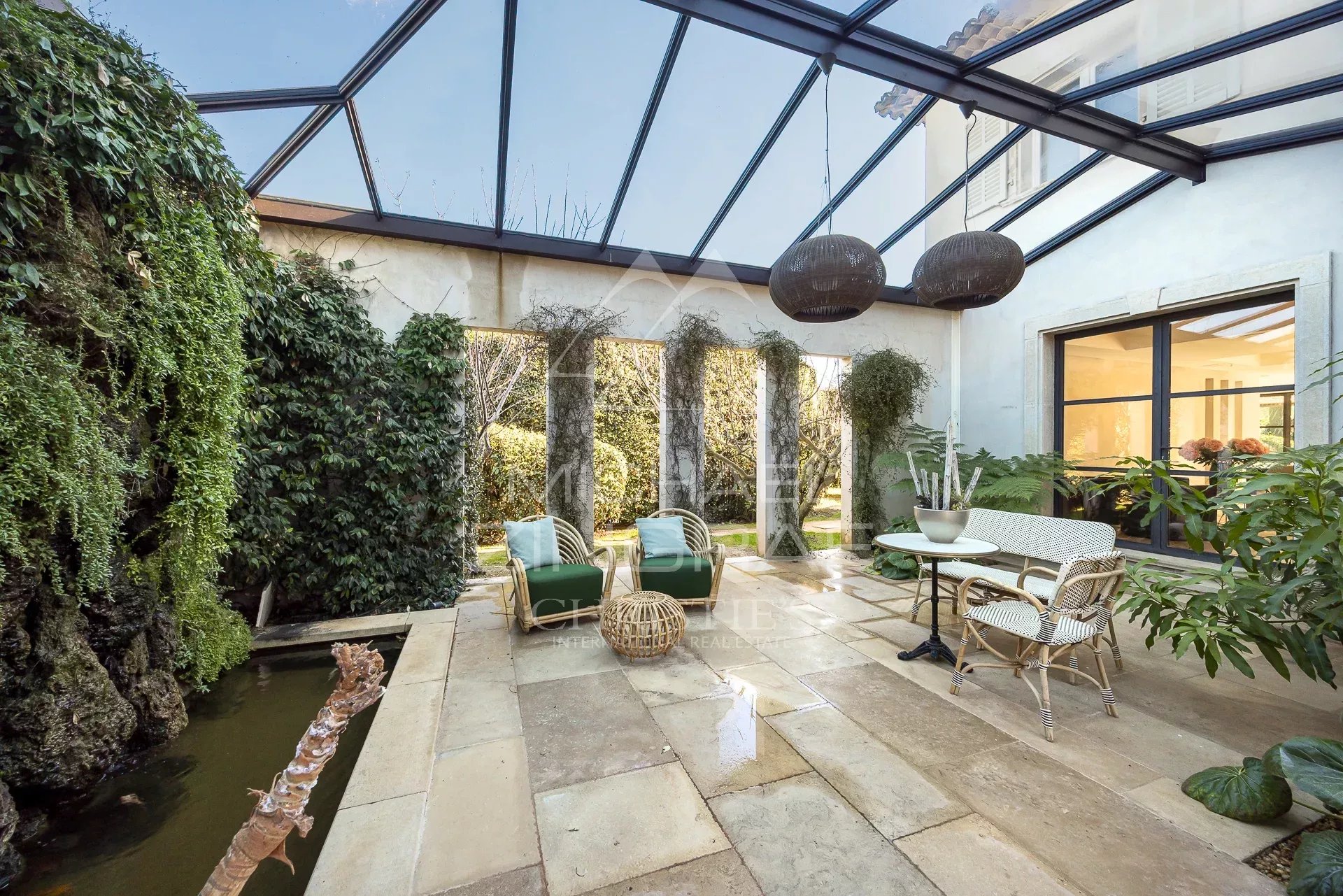 Glass-covered patio with wicker seating area and potted plants, overlooking a garden.