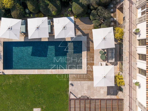 Aerial view of a rectangular swimming pool surrounded by white umbrellas and a wooden deck overlooking a green lawn.
