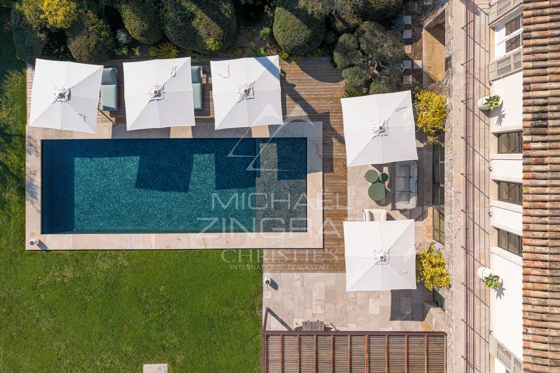Aerial view of a rectangular swimming pool surrounded by white umbrellas and a wooden deck overlooking a green lawn.