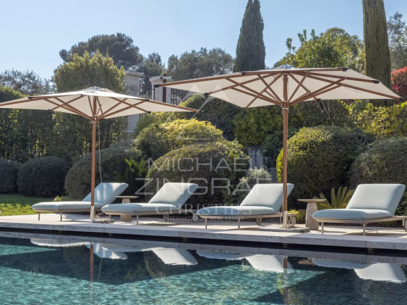 Poolside lounge chairs under large white umbrellas beside a tiled pool with manicured hedges in the background.