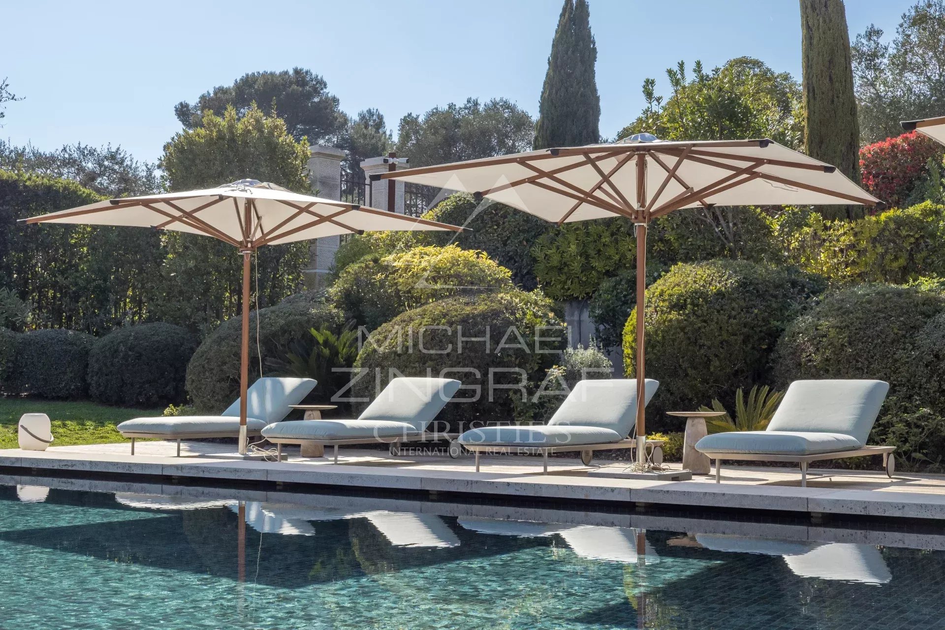 Poolside lounge chairs under large white umbrellas beside a tiled pool with manicured hedges in the background.