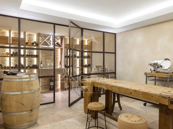 Wine cellar with glass-framed partitions, brick walls, and backlit wine shelves; a wooden barrel in the foreground and a rustic table beyond.