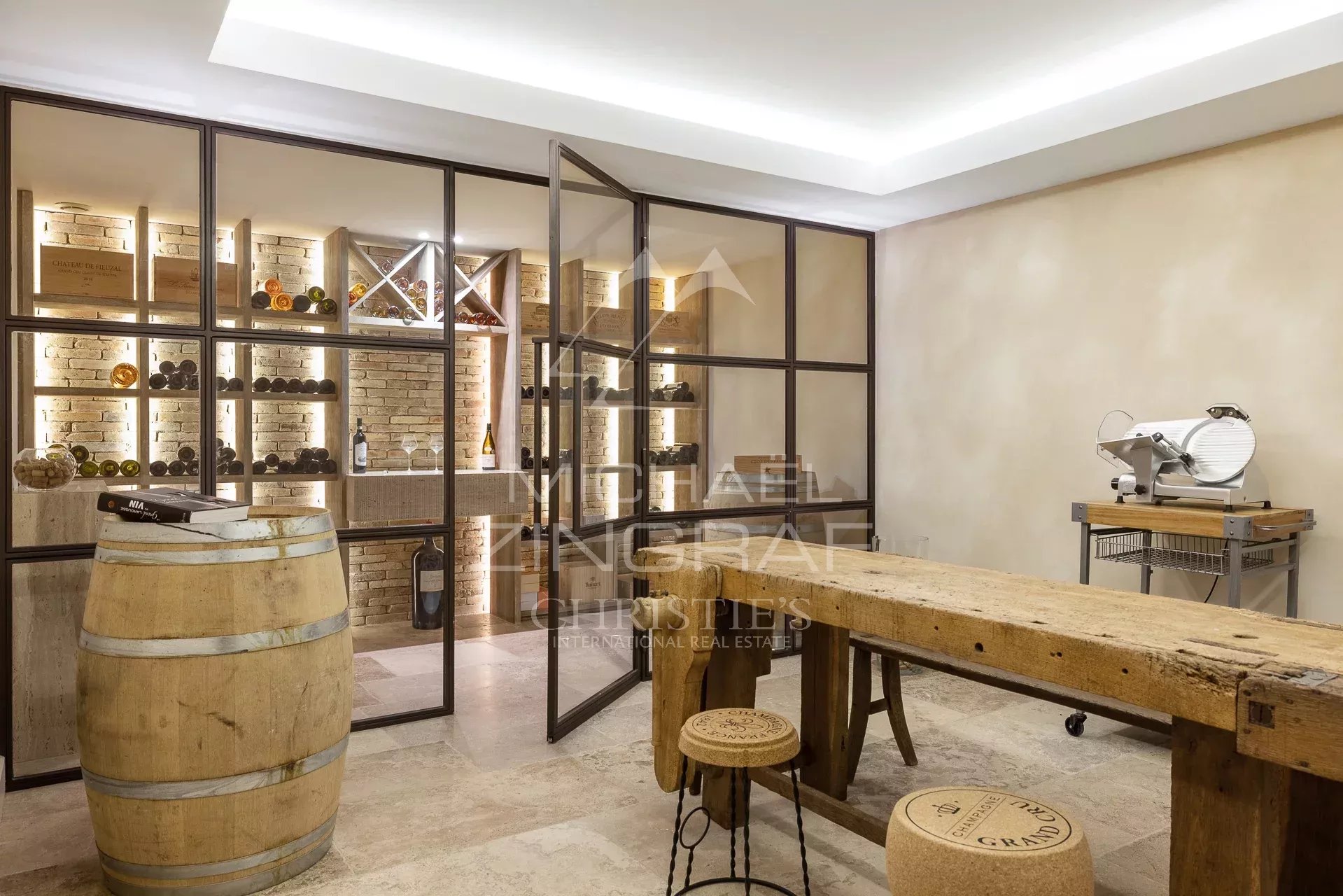 Wine cellar with glass-framed partitions, brick walls, and backlit wine shelves; a wooden barrel in the foreground and a rustic table beyond.