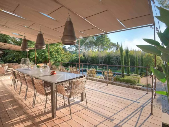 Wooden terrace with dining table and chairs, surrounded by greenery, near a swimming pool.