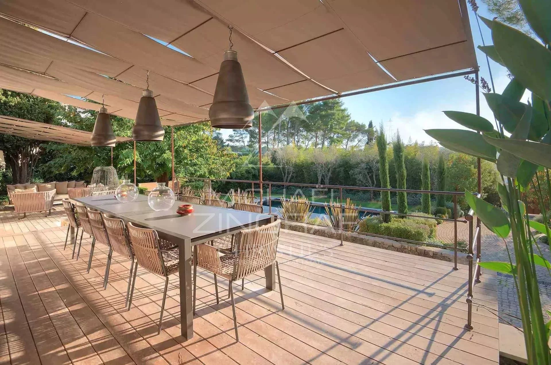 Terrace of a country house in Mougins Wooden terrace with dining table and chairs, surrounded by greenery, near a swimming pool.