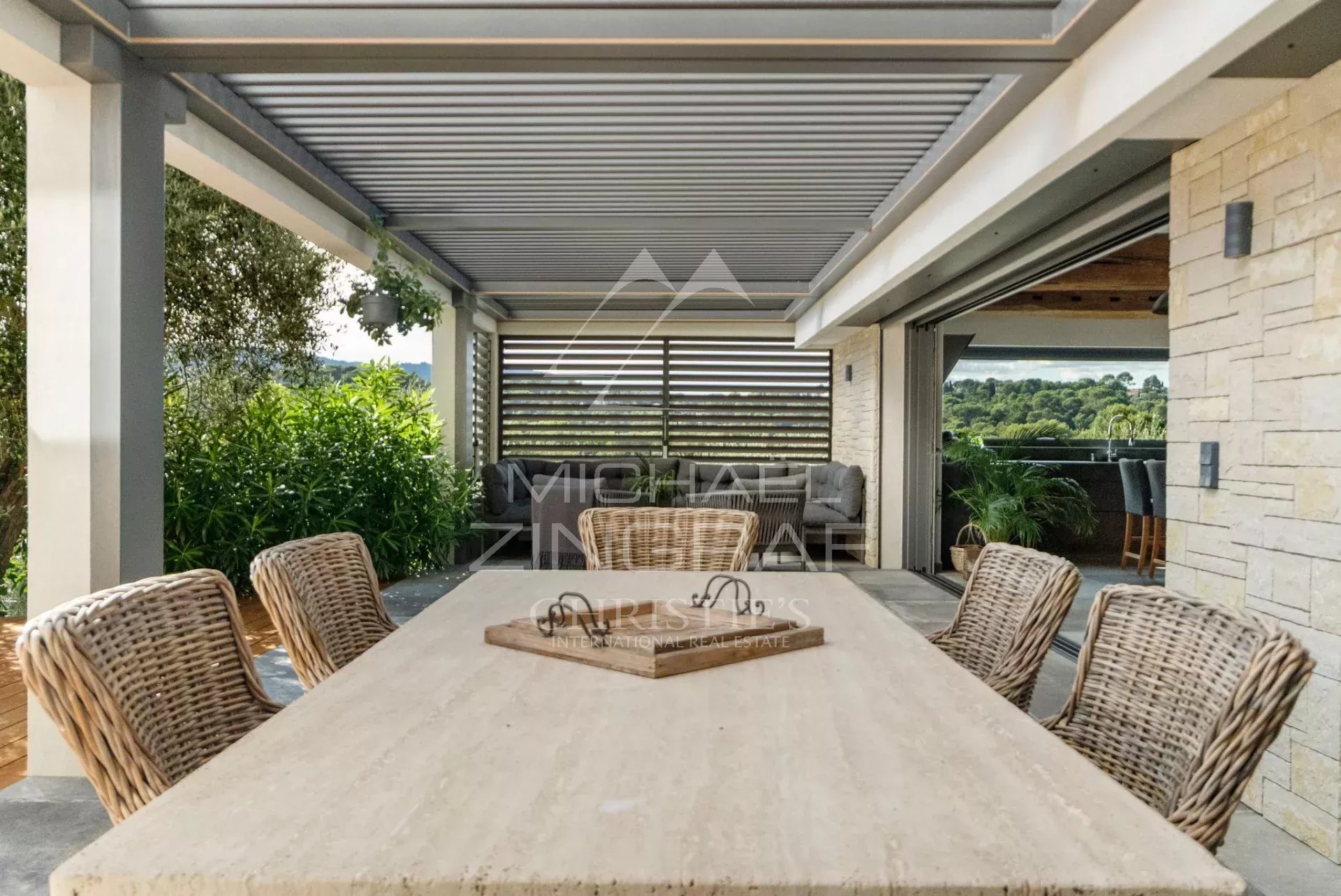 Terrasse d'une villa moderne avec table en pierre et chaises en osier, vue sur un environnement verdoyant.