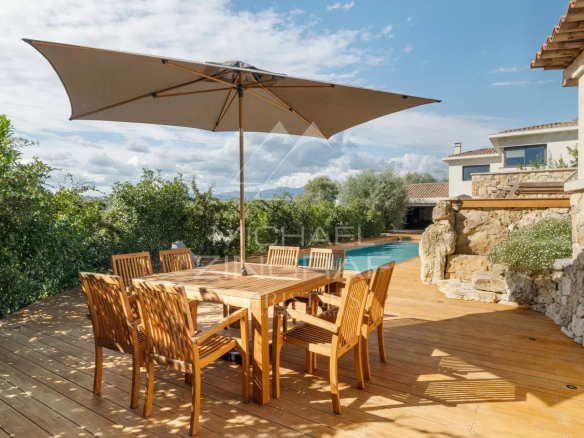 Terrasse en bois avec table et chaises en bois, parasol, piscine et verdure environnante.