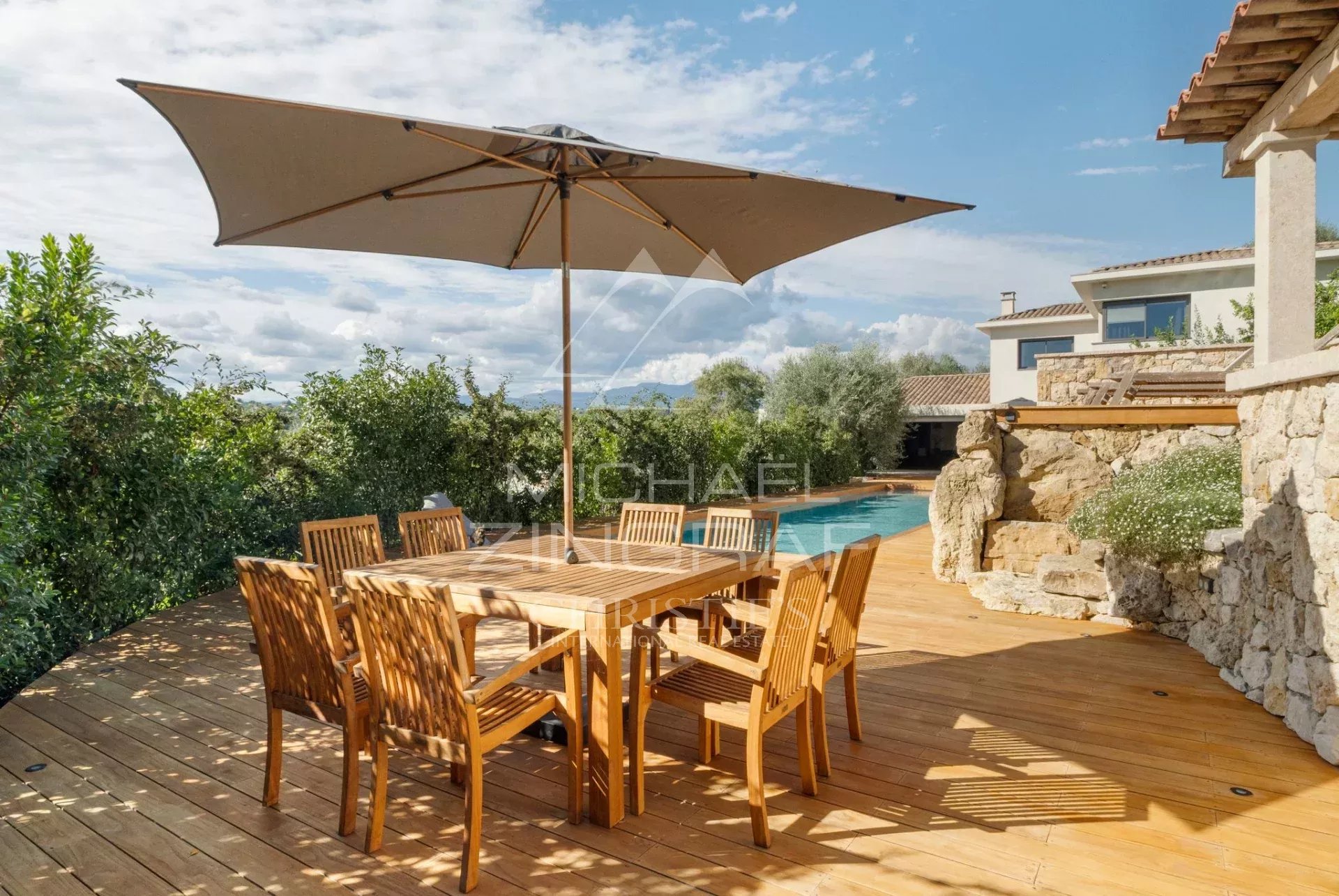 Terrasse en bois avec table et chaises en bois, parasol, piscine et verdure environnante.